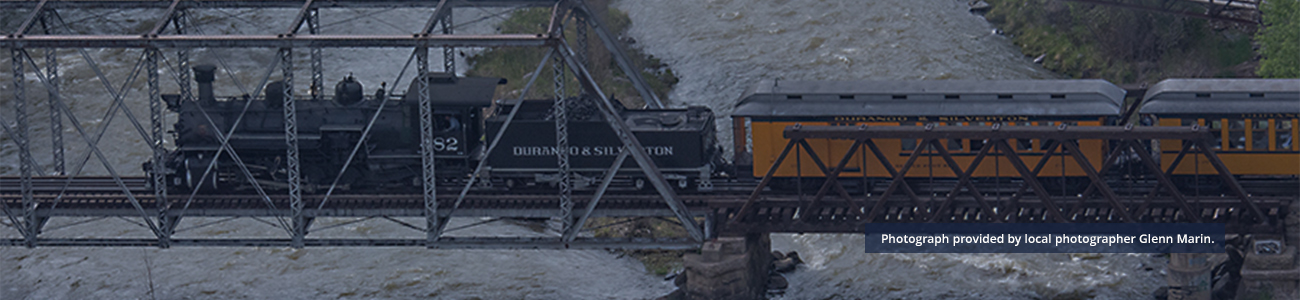 Vintage steam train from the Durango & Silverton line crossing a metal truss bridge over turbulent waters in Durango County. Image captured by Glenn Marin.
