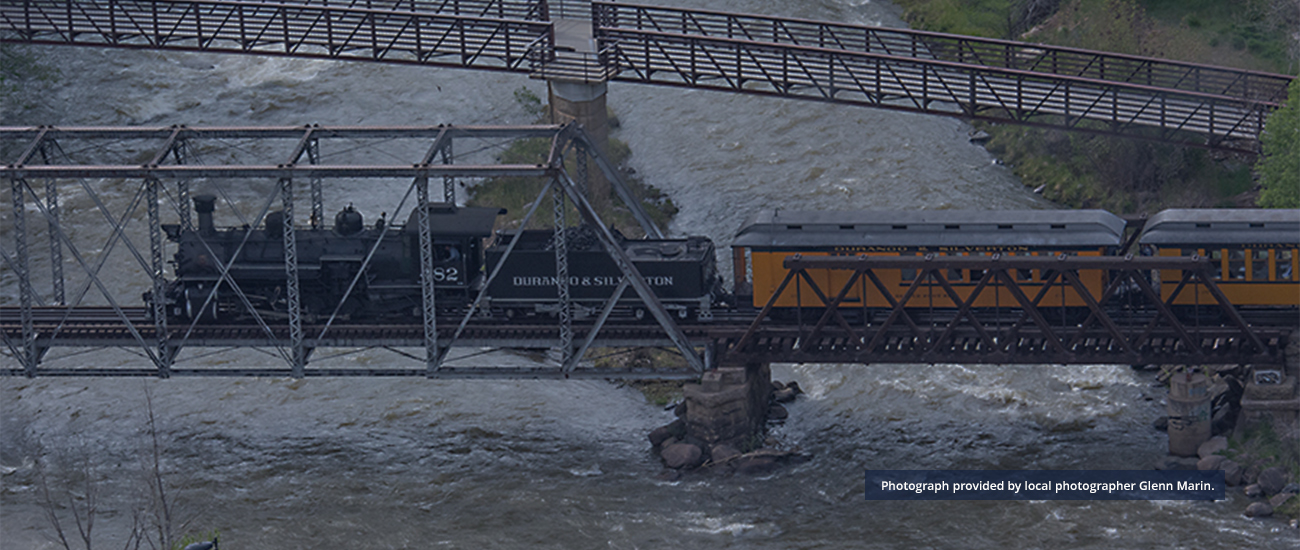 Vintage steam train from the Durango & Silverton line crossing a metal truss bridge over turbulent waters in Durango County. Image captured by Glenn Marin.